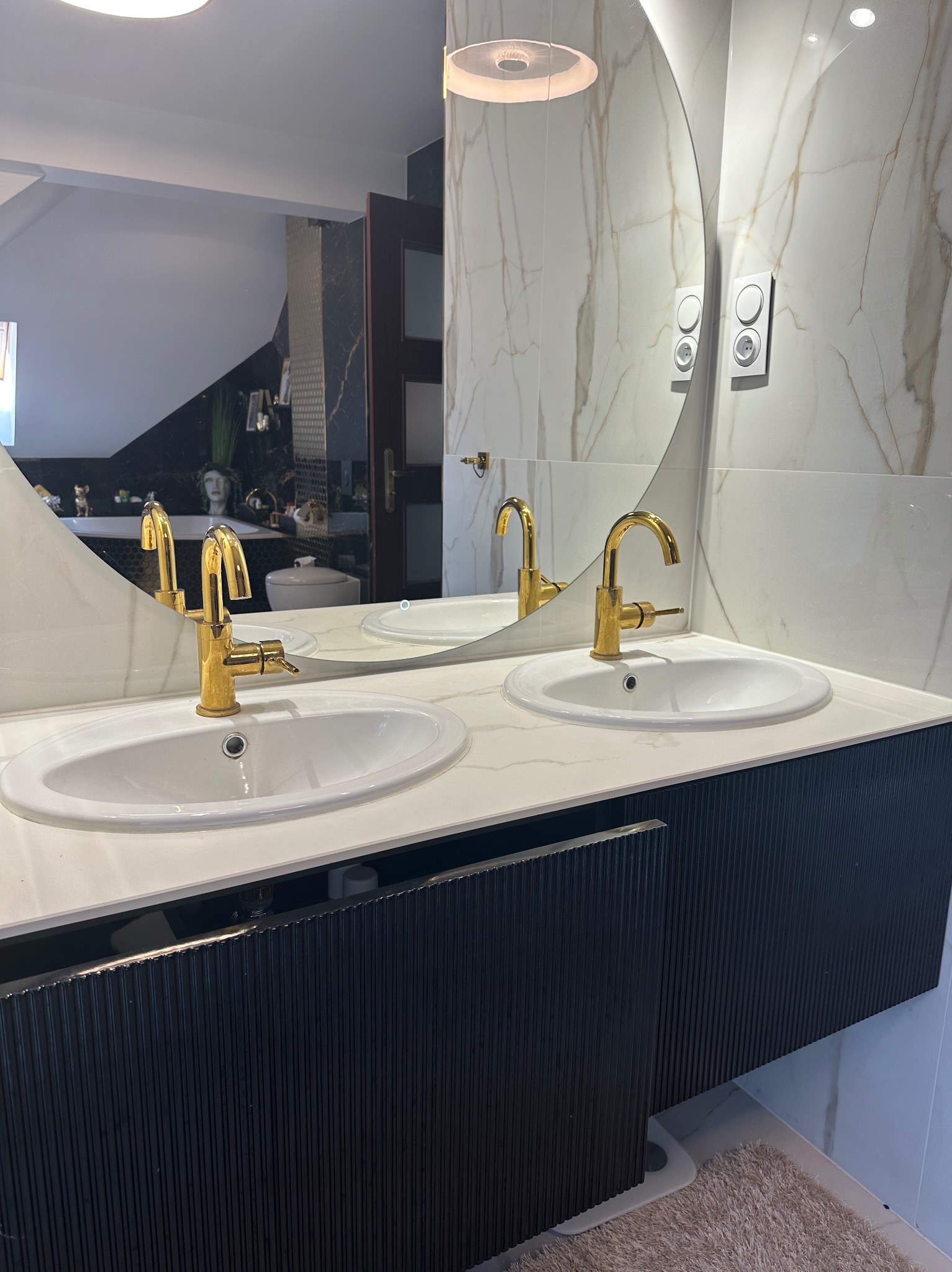 Nowoczesna łazienka z dwoma umywalkami, złotymi bateriami i okrągłym lustrem. Marmurowe ściany i ciemna szafka pod umywalką dodają elegancji.