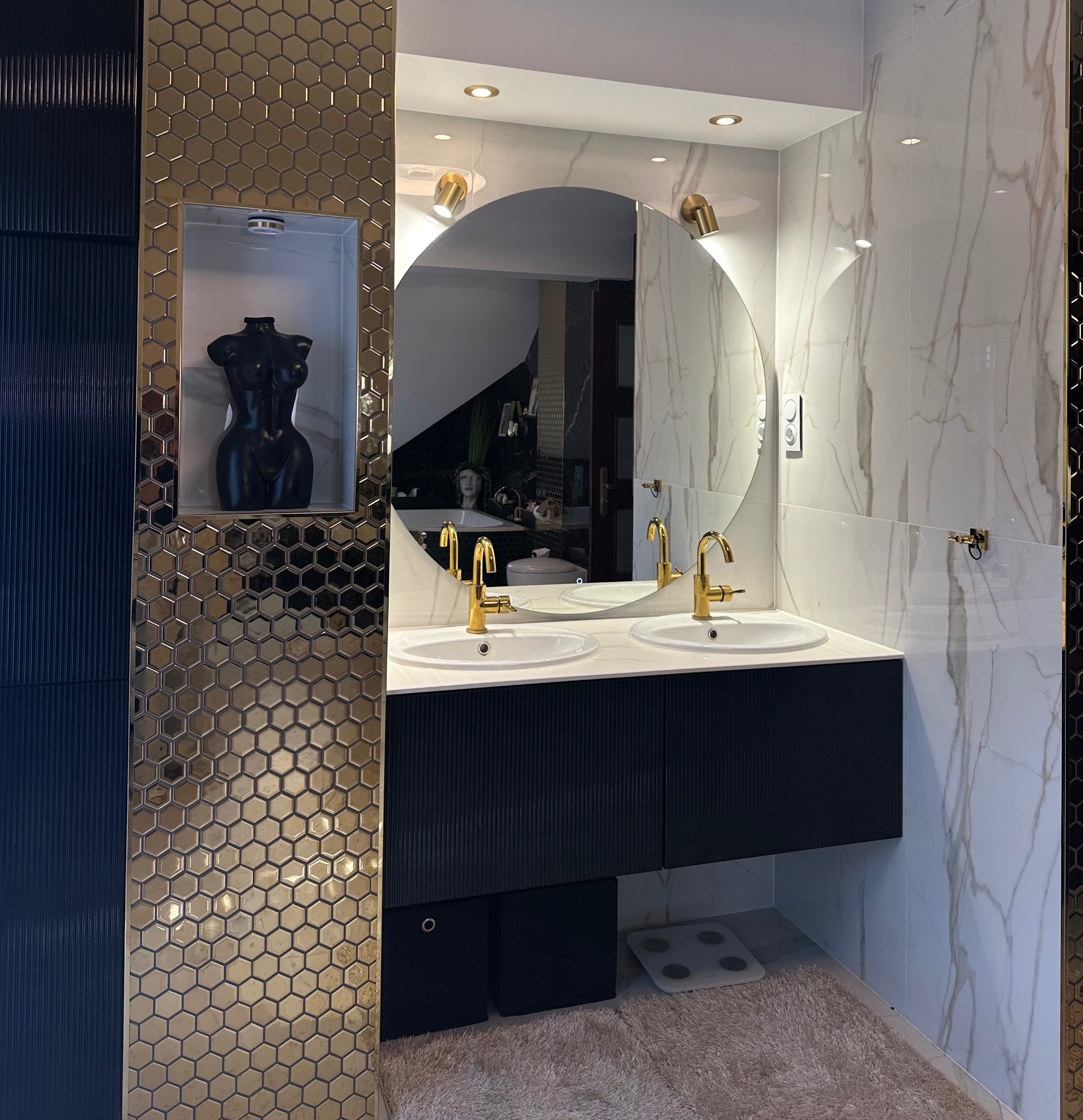 Nowoczesna łazienka z podwójną umywalką, złotymi bateriami i lustrem. Dekoracyjna mozaika i marmurowe płytki dodają elegancji. W tle widoczna rzeźba.