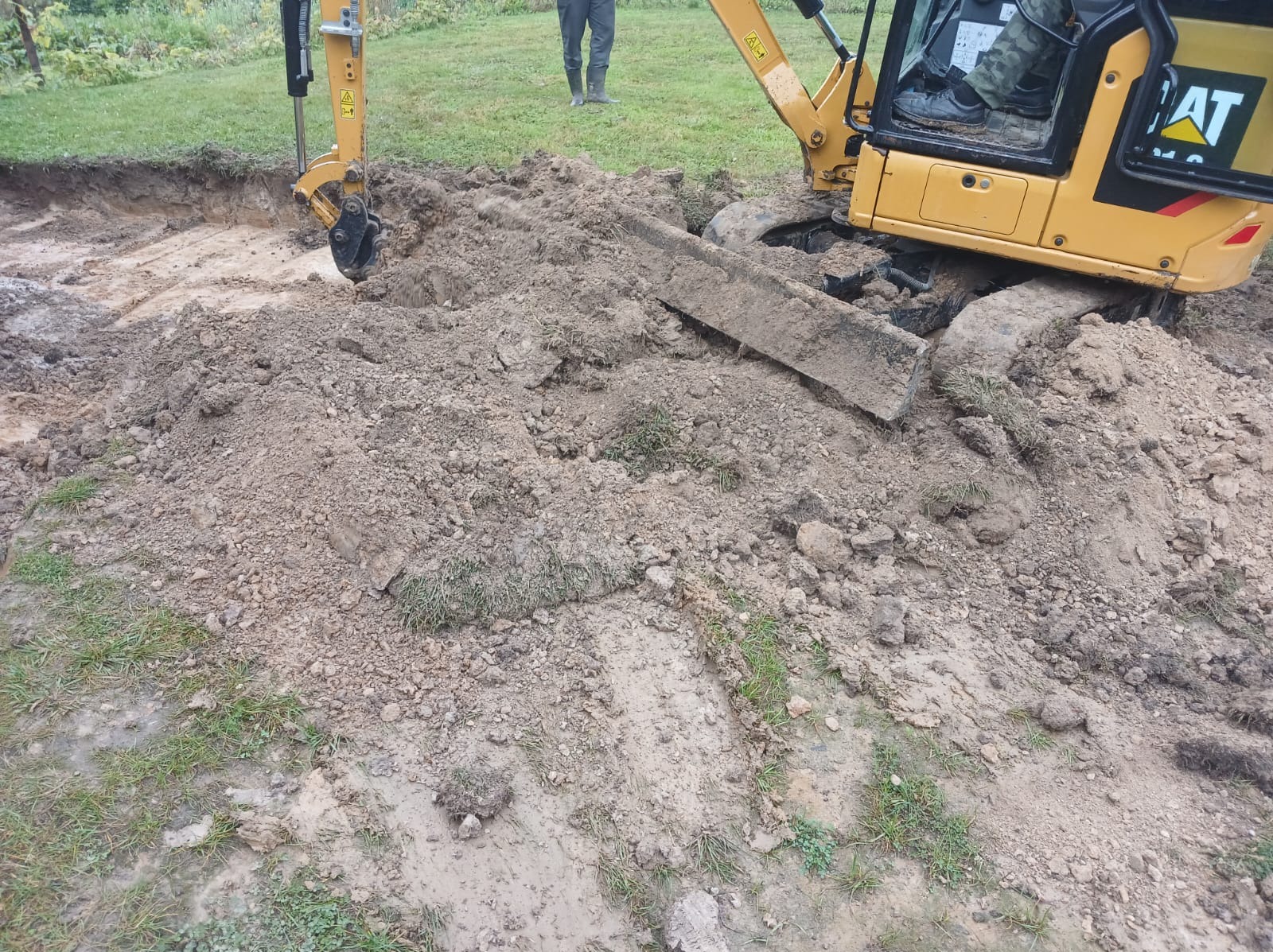 Żółta koparka CAT podczas wykopu ziemi w Nadziejowie. Widoczny fragment wykopu i operator w kabinie. Praca w toku, ziemia usypana wokół.