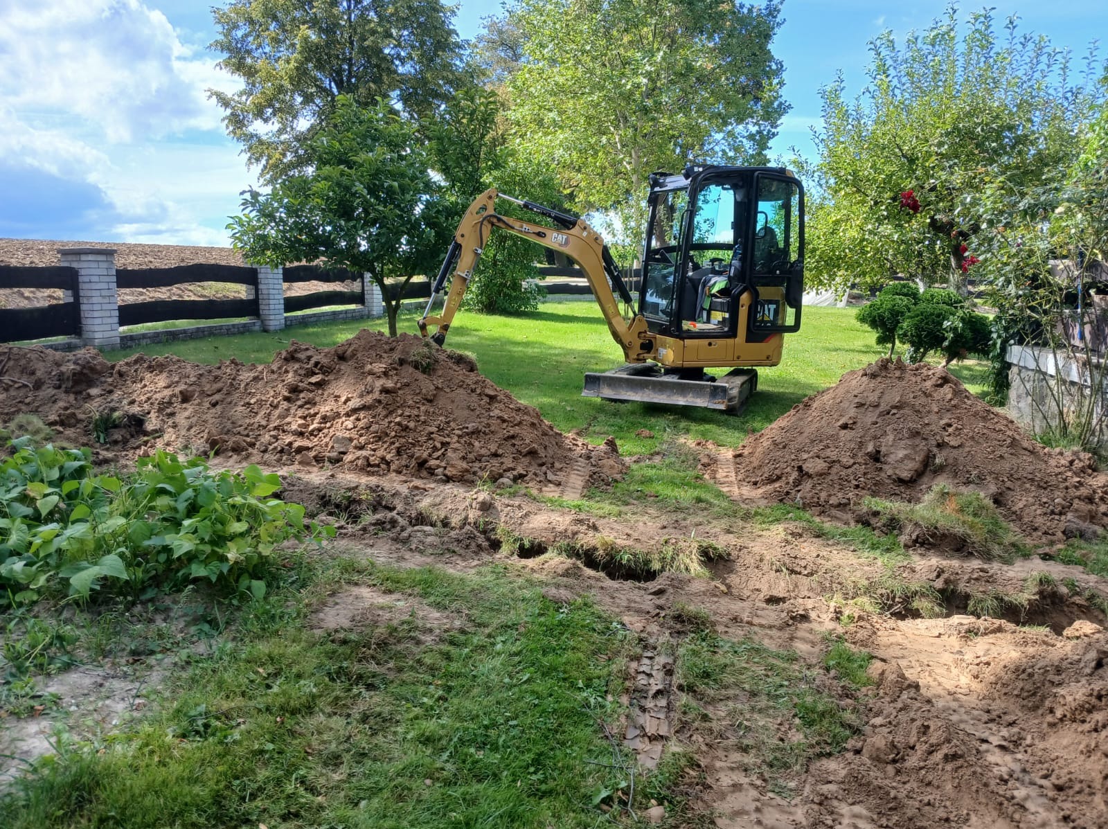 Żółta mini koparka CAT na trawniku, pomiędzy dwoma kopcami ziemi. Ślady opon na trawie. W tle ogrodzenie i drzewa.