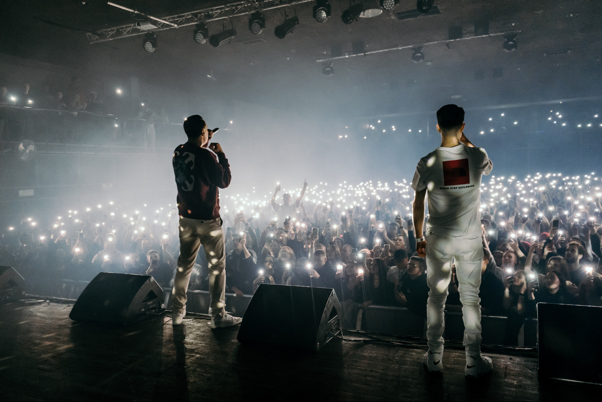 Dwóch artystów na scenie, oświetlonych światłami reflektorów, występuje przed tłumem fanów, którzy trzymają w górze telefony z włączonymi latarkami, tworząc morze świateł.