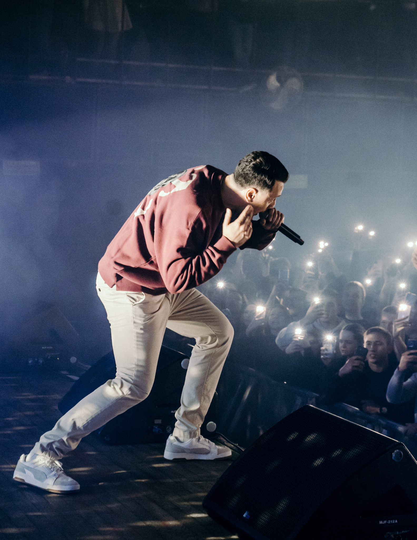 Artysta w bordowej bluzie i jasnych spodniach rapuje do mikrofonu na scenie oświetlonej reflektorami, w tle widownia z telefonami.