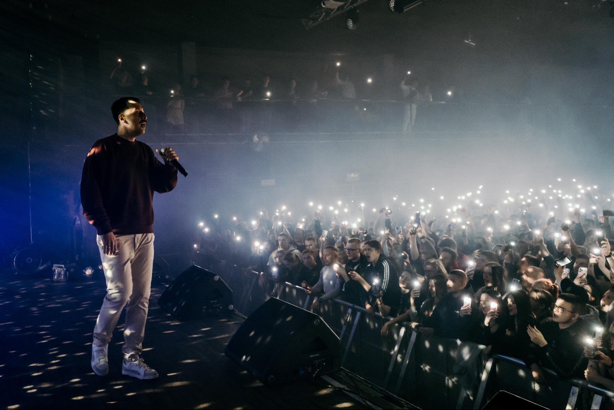 Wokalista na scenie, trzyma mikrofon podczas koncertu z tłumem fanów w tle, którzy oświetlają go telefonami. Scena oświetlona reflektorami.