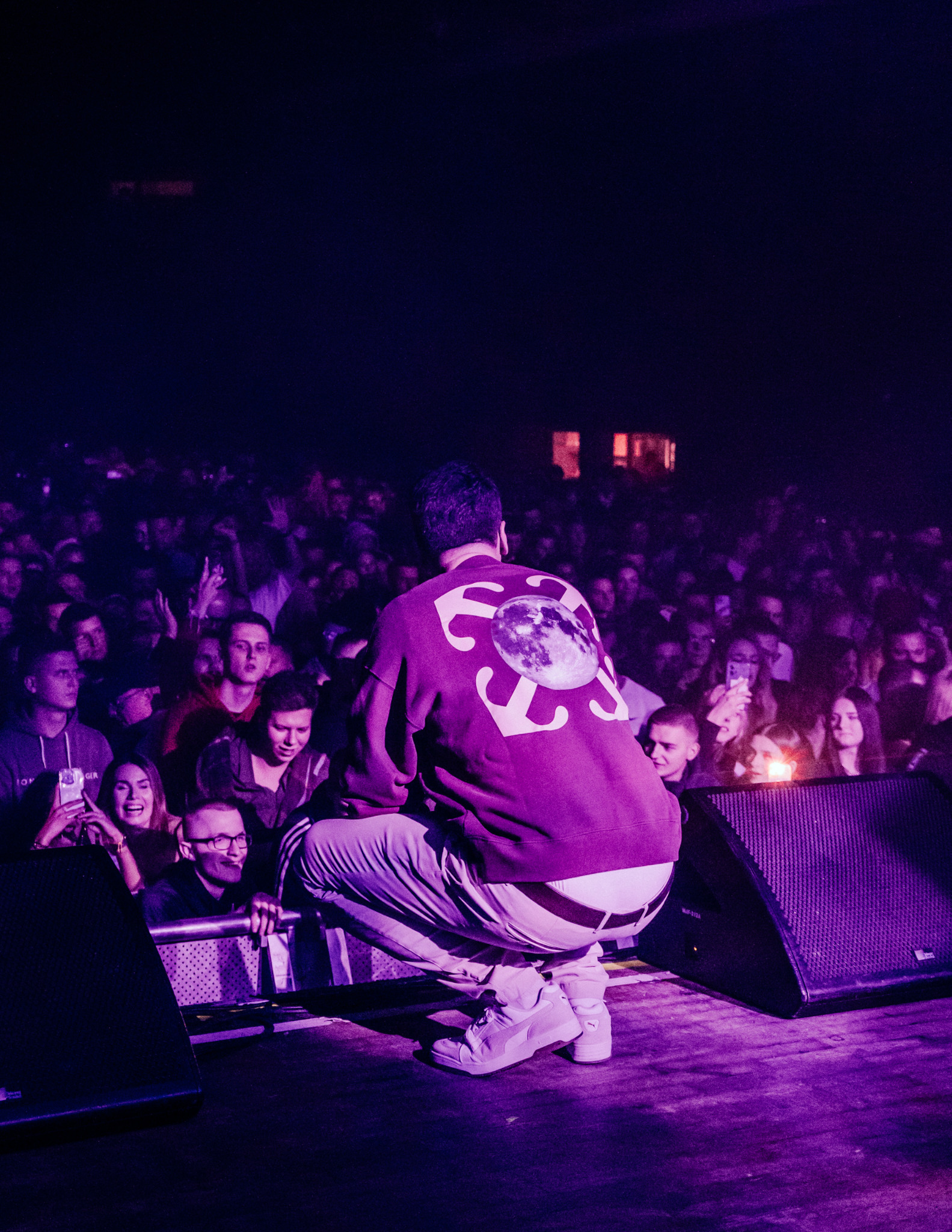 Artysta na scenie, widziany od tyłu, w fioletowym oświetleniu. W tle tłum ludzi i głośniki. Na bluzie duży nadruk z motywem księżyca i strzałek.