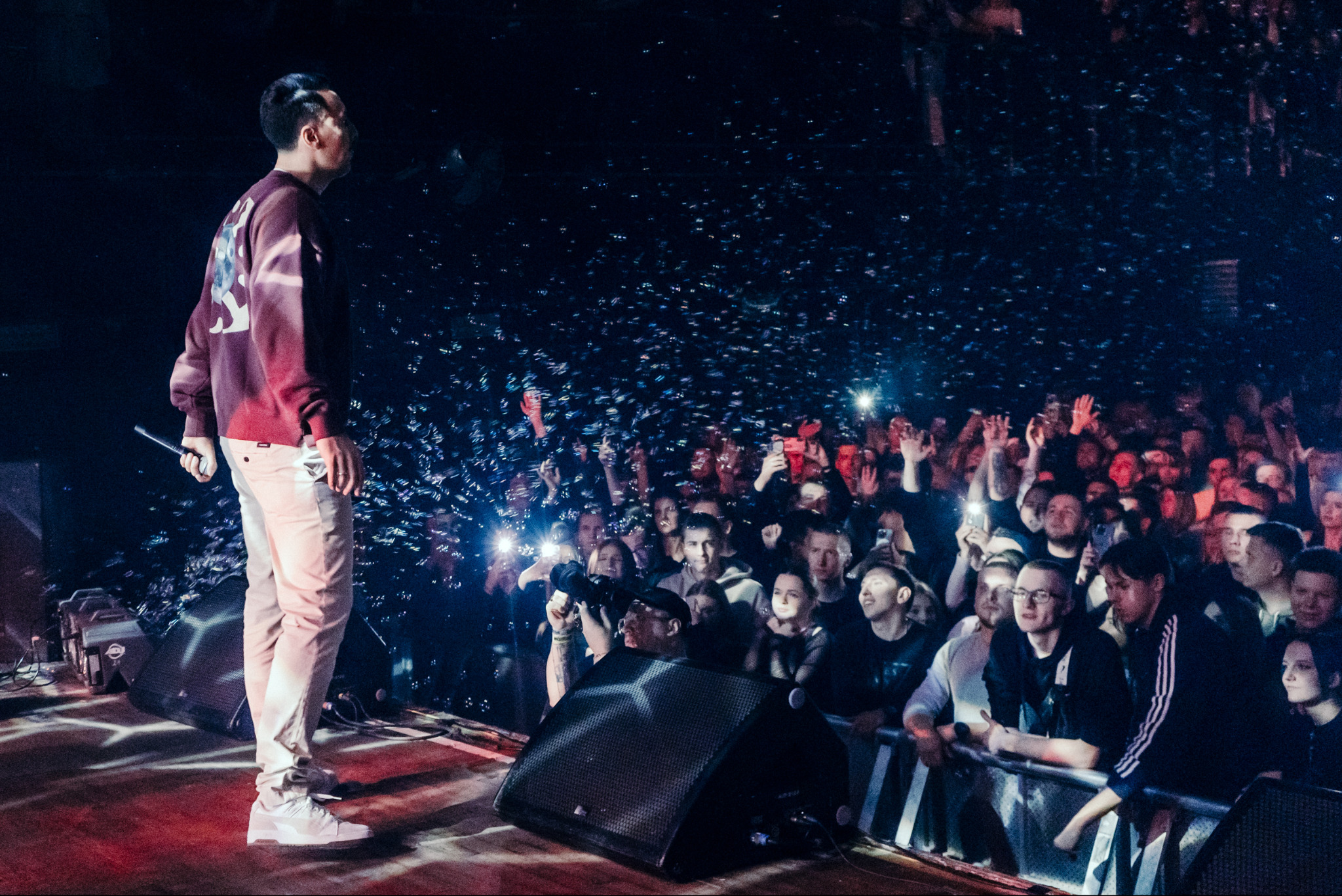 Piosenkarz na scenie, oświetlony światłami, z mikrofonem w dłoni, zwrócony do publiczności z podniesionymi rękami i telefonami. Koncert w ciemnym otoczeniu.