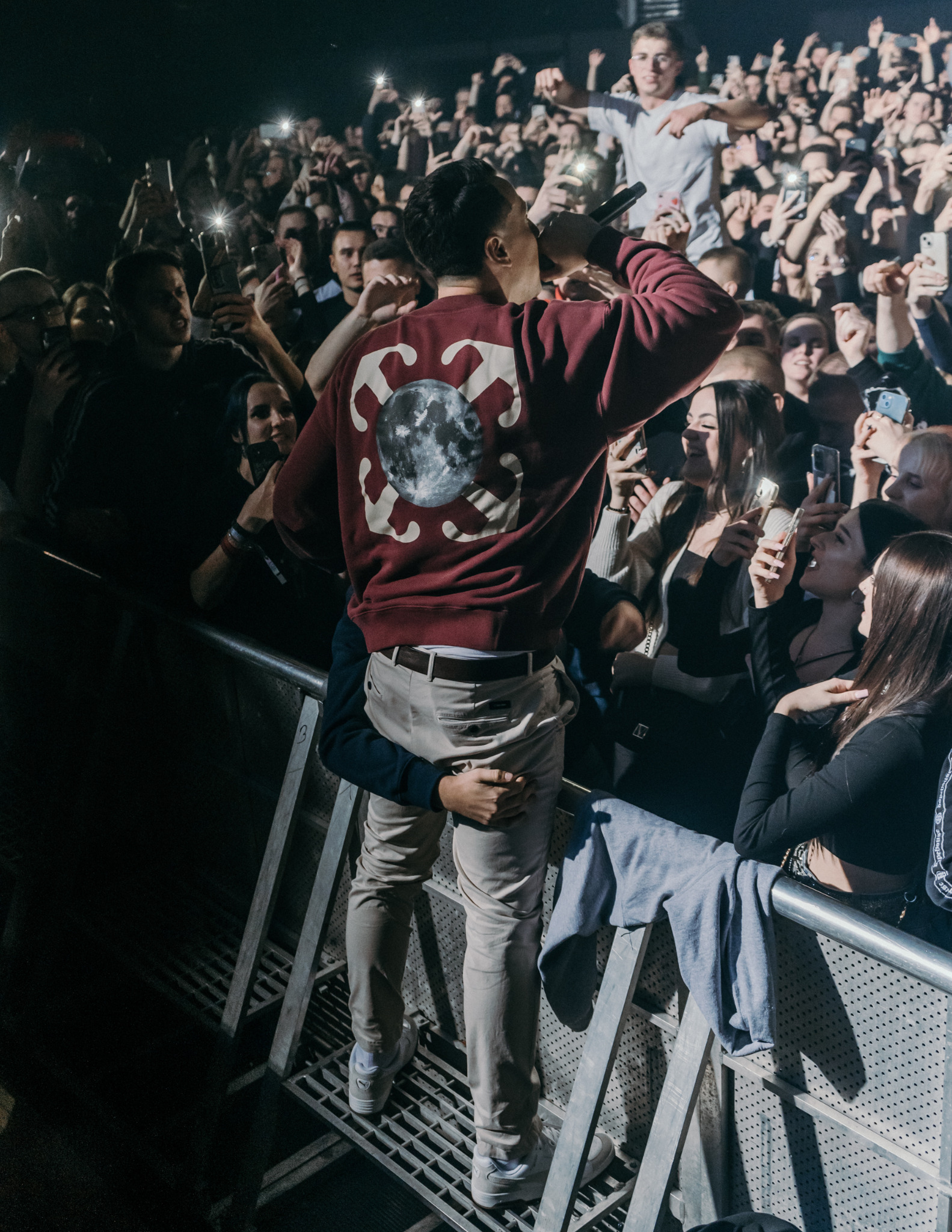 Koncert muzyczny: piosenkarz w bordowej bluzie z mikrofonem na scenie, tłum fanów z telefonami w tle. Ujęcie z dołu, dynamiczna atmosfera.