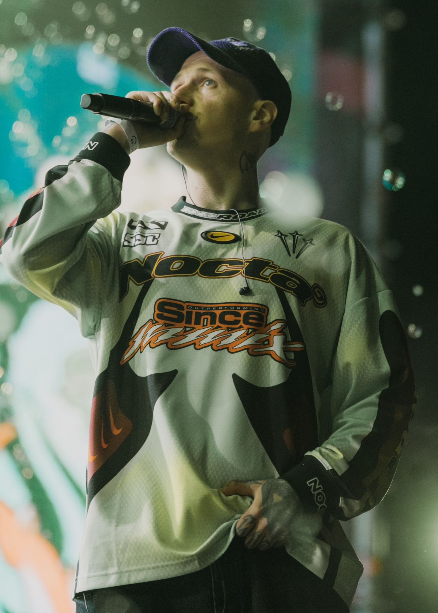 Młody mężczyzna z tatuażami, w bluzie z długim rękawem i czapce z daszkiem, śpiewa do mikrofonu na tle rozmytej sceny.