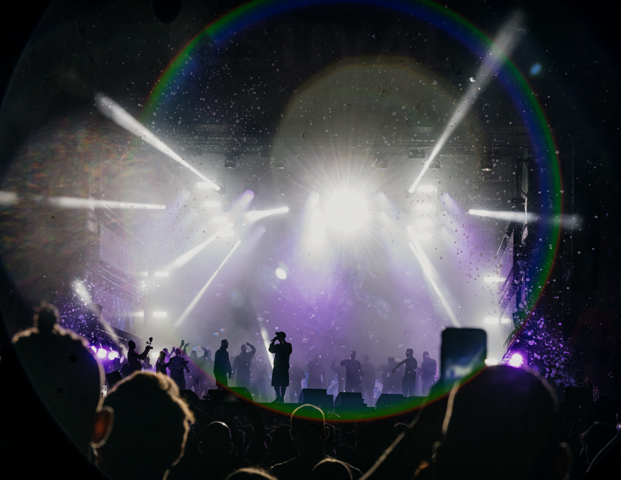 Rozświetlona scena koncertowa z silnymi reflektorami i sylwetkami artystów na tle tłumu. Widoczny efekt tęczy na obiektywie i unoszące się drobinki w powietrzu.