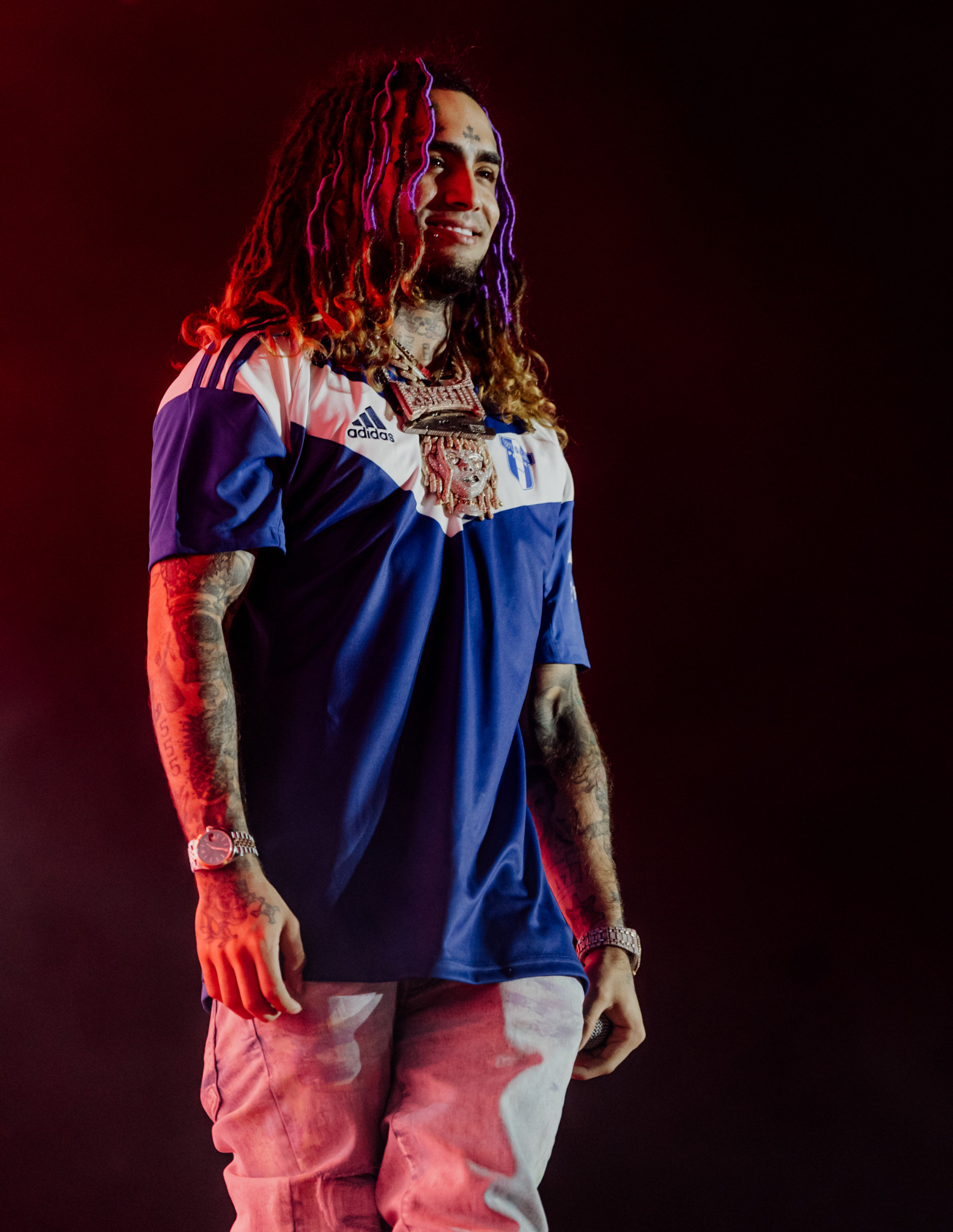 Młody raper z dredami i tatuażami, w sportowej koszulce Adidas i z ciężkim łańcuchem, uśmiecha się na scenie oświetlonej czerwonym światłem.