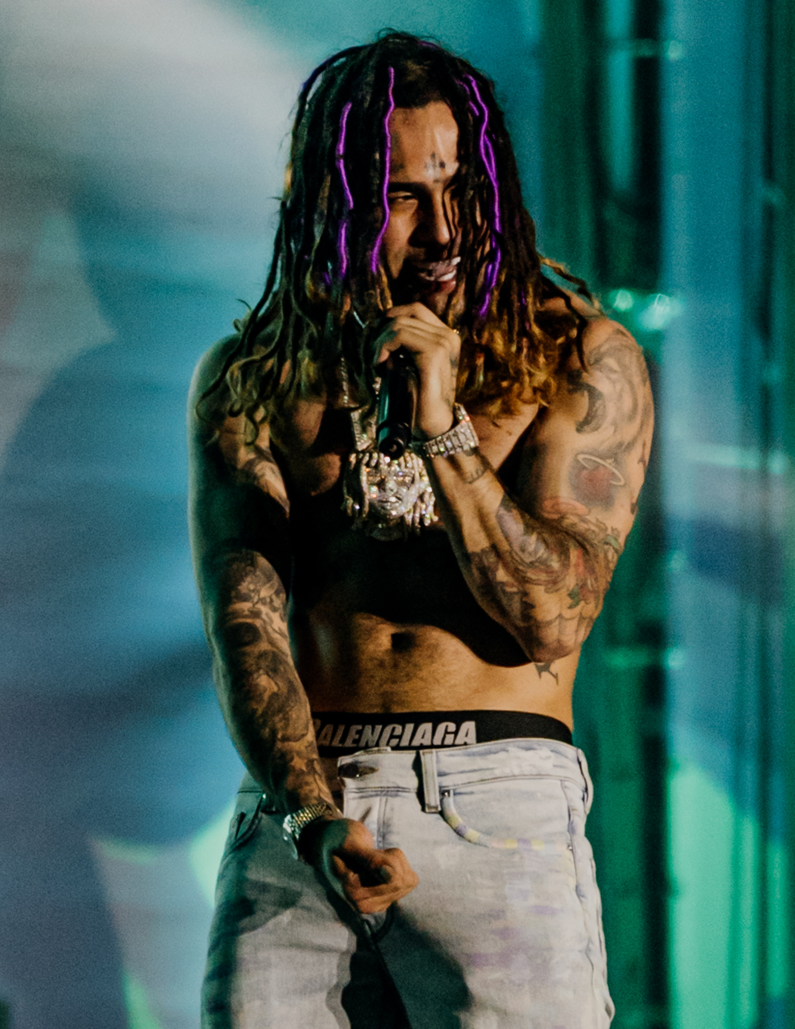 Mężczyzna z dredami i tatuażami śpiewa do mikrofonu na scenie, ubrany w dżinsy Balenciaga i biżuterię. Scena oświetlona jest zielonym i niebieskim światłem.