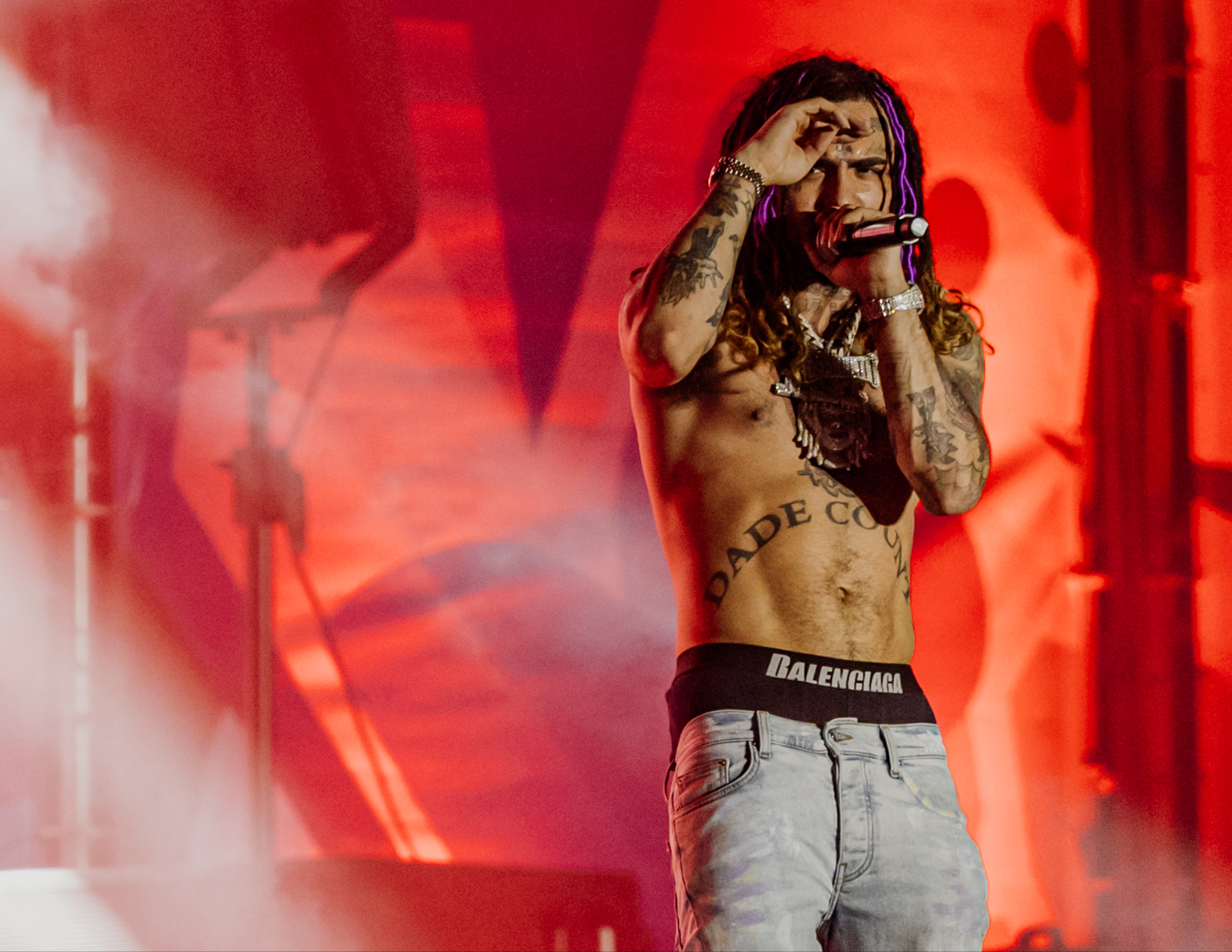 Raper z dredami i tatuażami na scenie w czerwonym oświetleniu, w trakcie występu. Mikrofon w dłoni, robi gest dłonią nad okiem. Ubrany w dżinsy i markową bieliznę.