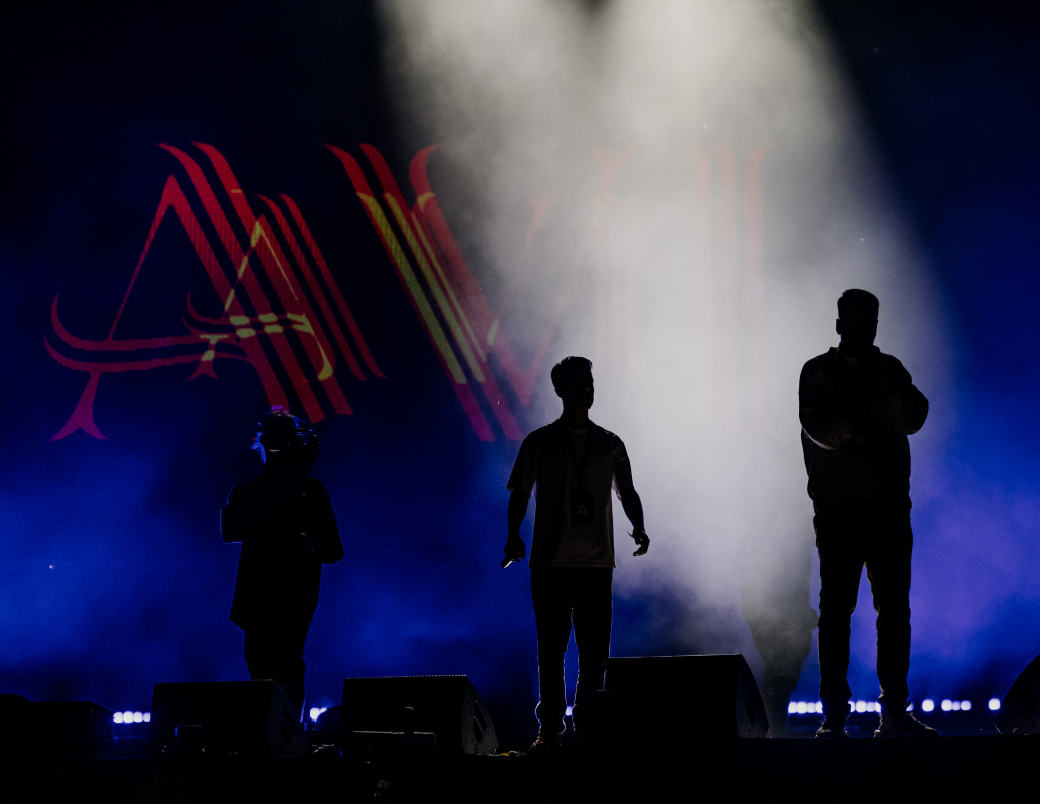 Silhouetted figures on stage at a Warszawa event, bathed in blue and white light with a large logo projected behind them. Dynamic stage photography.