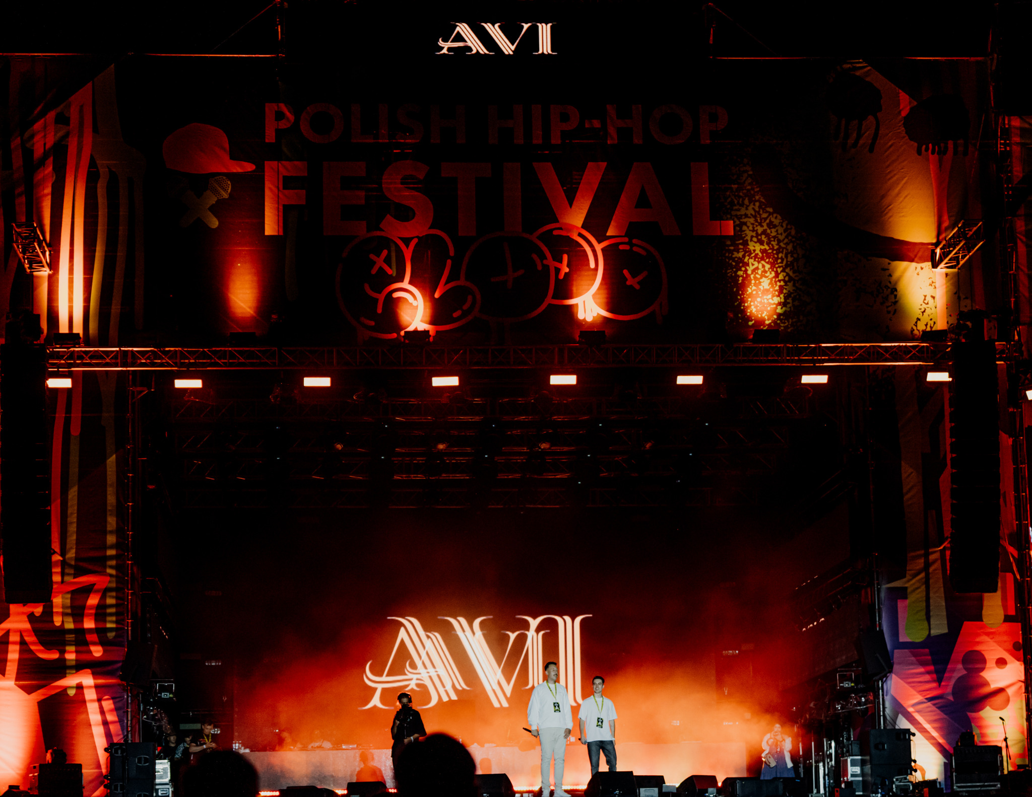 Zdjęcie z festiwalu Polish Hip-Hop z artystami na scenie AVI, oświetlonej czerwonymi światłami. W tle baner z nazwą festiwalu i graffiti.