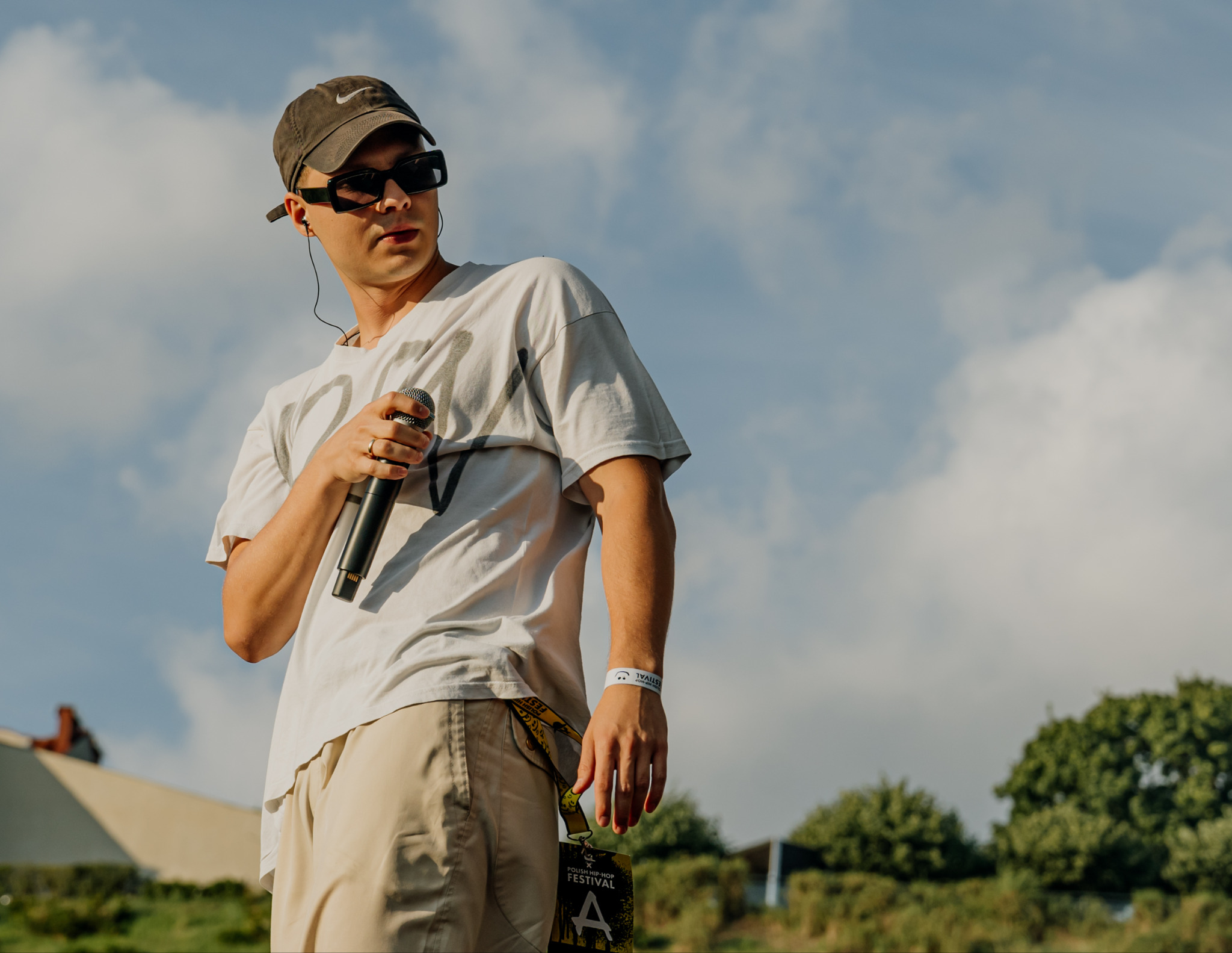 Młody mężczyzna w okularach i czapce, trzymający mikrofon na tle nieba z chmurami, z identyfikatorem festiwalowym, sugerujący występ artystyczny.
