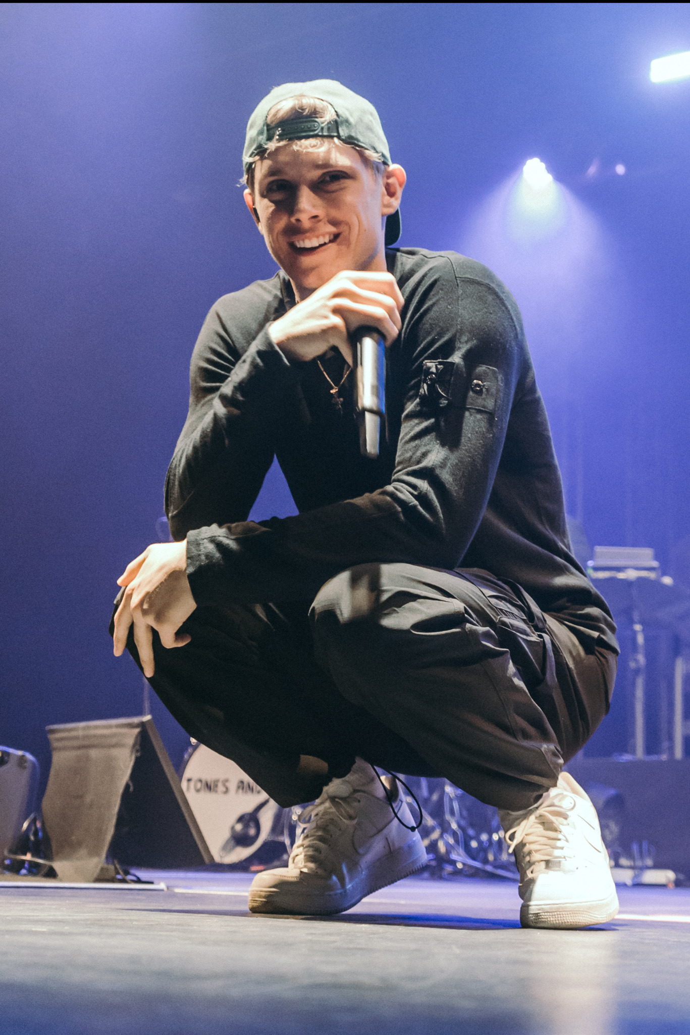 Uśmiechnięty mężczyzna w kucki na scenie, trzyma mikrofon. W tle instrumenty muzyczne i niebieskie oświetlenie. Ubrany w czarne spodnie, bluzę i białe buty sportowe.