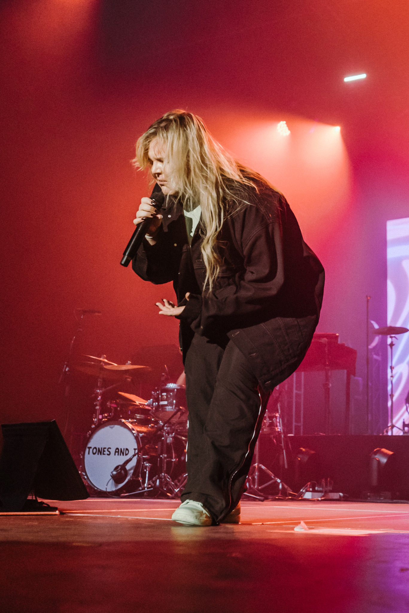 Wokalistka Tones and I w trakcie występu na scenie, w dynamicznej pozie z mikrofonem, w tle perkusja i czerwono-różowe oświetlenie sceniczne.
