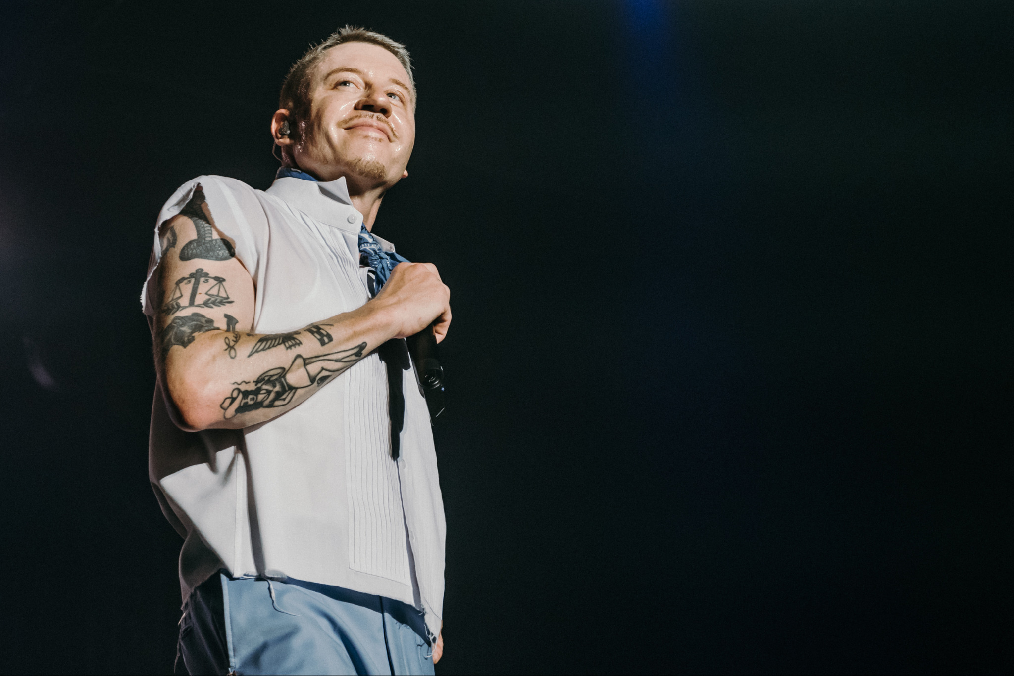 Mężczyzna z tatuażami na ramieniu, w jasnej koszuli i niebieskiej apaszce, trzyma mikrofon na scenie w ciemnym otoczeniu. Uśmiechnięty, patrzy w górę.