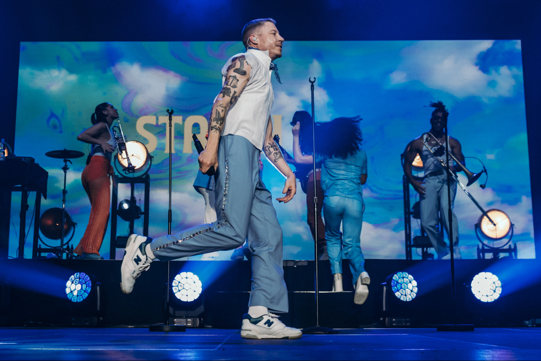 Dynamiczne ujęcie muzyka z tatuażami na scenie podczas występu na tle ekranu z motywem chmur i niebieskim oświetleniem. Wokół niego zespół i instrumenty.