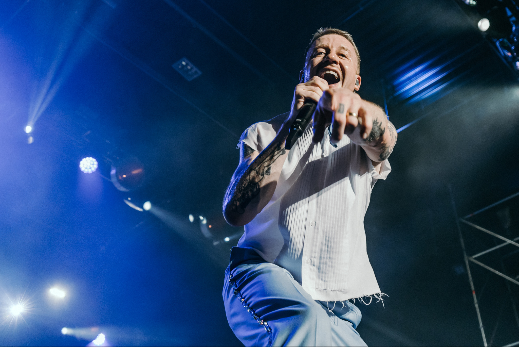 Energiczny wokalista z tatuażami, w białej koszuli, śpiewa do mikrofonu na scenie oświetlonej niebieskimi światłami, wskazując palcem na widownię.