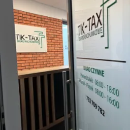 Wejście do biura rachunkowego TIK-TAX z godzinami otwarcia na szklanych drzwiach. W tle logo firmy na ścianie imitującej cegłę.