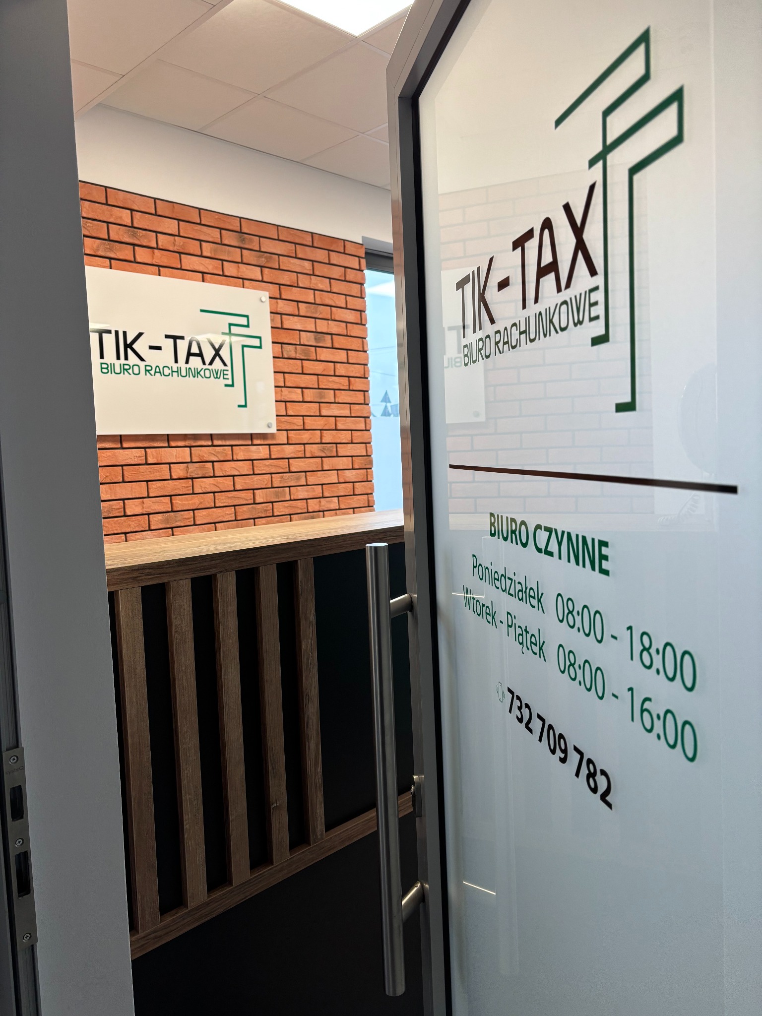 Wejście do biura rachunkowego TIK-TAX z godzinami otwarcia na szklanych drzwiach. W tle logo firmy na ścianie imitującej cegłę.