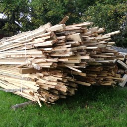 TARTAK DOBCZYCE Michał Knapik - Stos tarcicy budowlanej związany sznurkiem, ułożony na trawie. W tle drzewa i budynek. Deski o nieregularnych kształtach, jasne drewno, widoczne sęki.