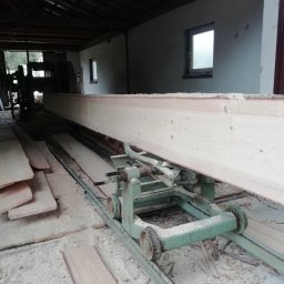 TARTAK DOBCZYCE Michał Knapik - Wnętrze tartaku: duża, jasna belka tarcicy na zielonym wózku transportowym, w tle maszyny i deski, wszystko pokryte trocinami. Widoczne okna w tle.