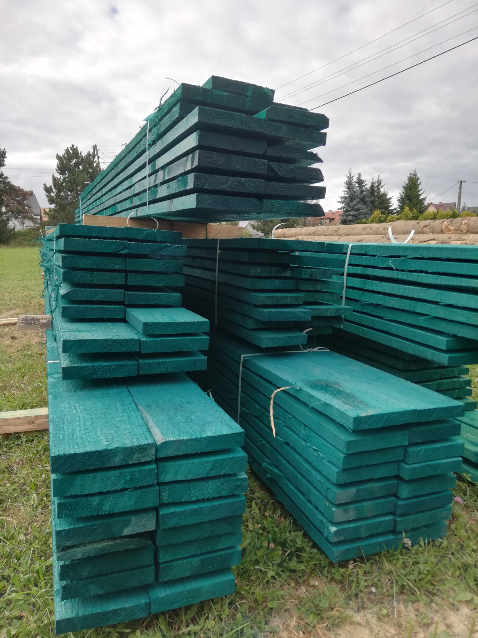 Świeżo pomalowana na zielono tarcica budowlana ułożona w stosy, zabezpieczona sznurkiem, na trawie w Dobczycach. Widok z dołu.