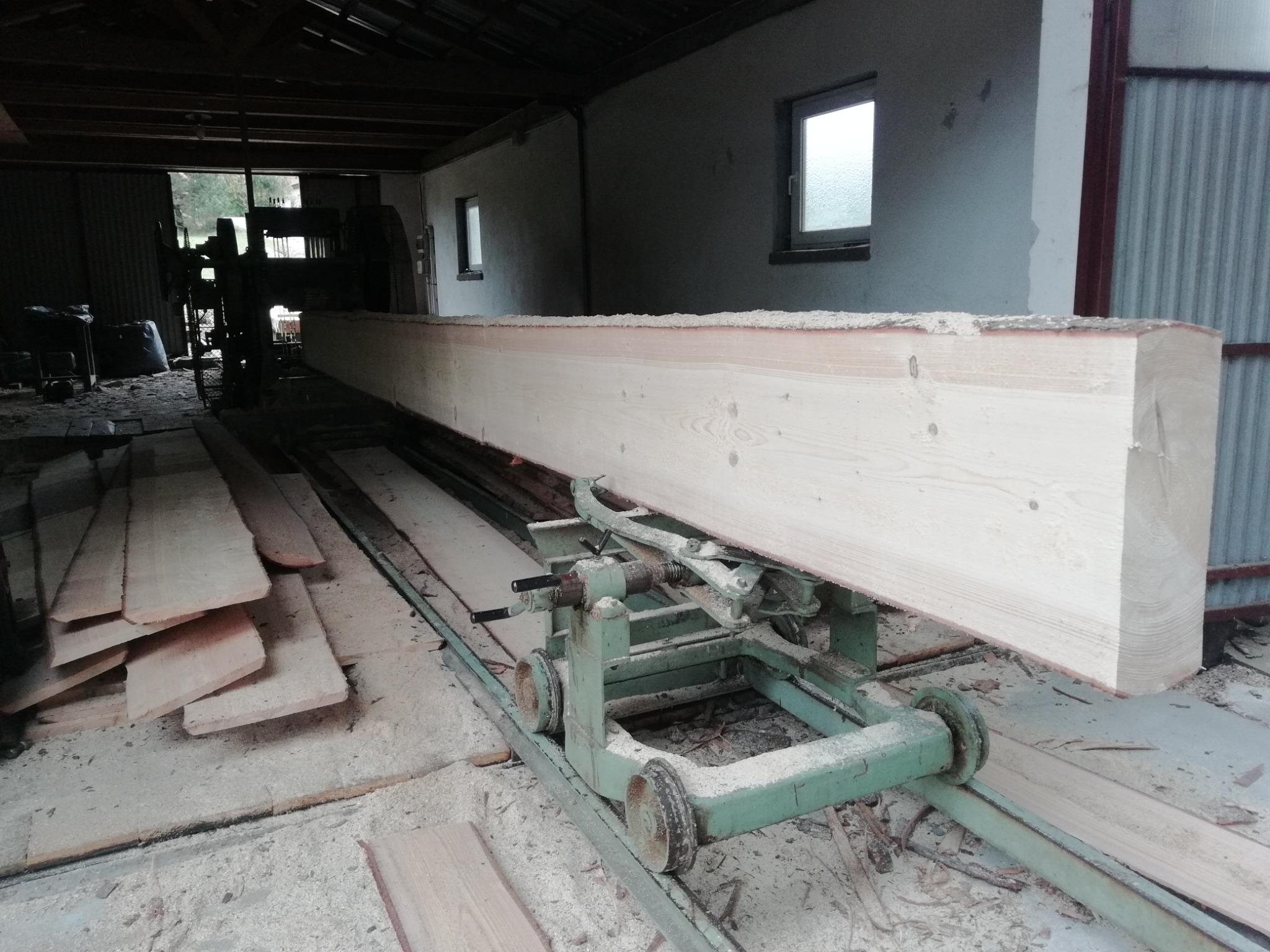 Wnętrze tartaku: duża, jasna belka tarcicy na zielonym wózku transportowym, w tle maszyny i deski, wszystko pokryte trocinami. Widoczne okna w tle.