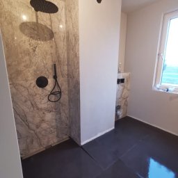 Karo - Wykończona łazienka z marmurowymi płytkami pod prysznicem i czarną podłogą. Widoczna armatura w kolorze czarnym i okno z widokiem na zewnątrz.