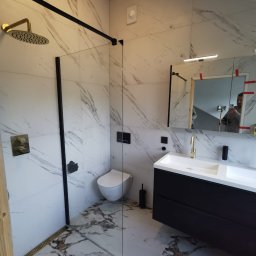 Karo - Nowoczesna łazienka z marmurowymi płytkami, czarnymi akcentami i złotą armaturą. Kabina prysznicowa typu walk-in z czarną ramą. Umywalka z szafką w kolorze czarnym.