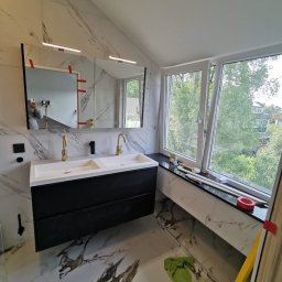 Karo - Wykończona łazienka z marmurowymi płytkami, czarną szafką pod umywalką z dwiema białymi umywalkami i złotą armaturą. Widok na zieleń za oknem.