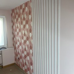 Karo - Ściana w pokoju po remoncie: dekoracyjna tapeta w geometryczne wzory i pionowe, białe lamele. Podłoga zabezpieczona kartonem.