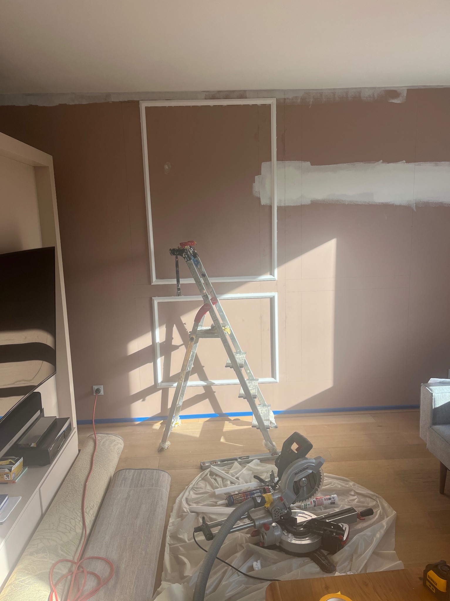 Pokój w trakcie dekoracji ścian: brązowe ściany z białymi listwami, drabina, piła do drewna i narzędzia na podłodze. Widoczne ślady malowania na górze ściany.