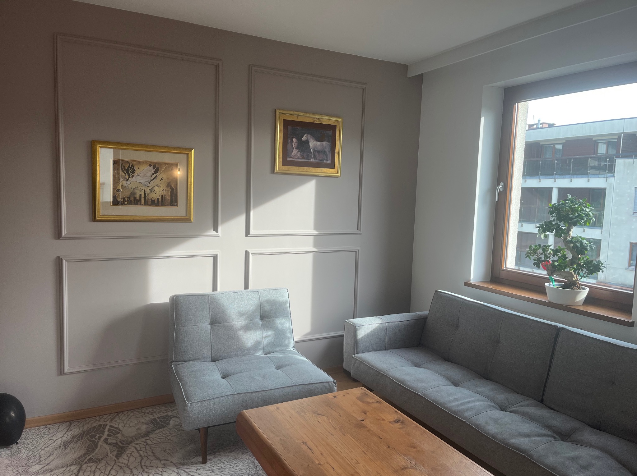 Jasne wnętrze salonu z szarą sofą, fotelem i drewnianym stolikiem kawowym. Na ścianie dekoracyjne ramy i obrazy w złotych ramach. W oknie widoczny budynek.