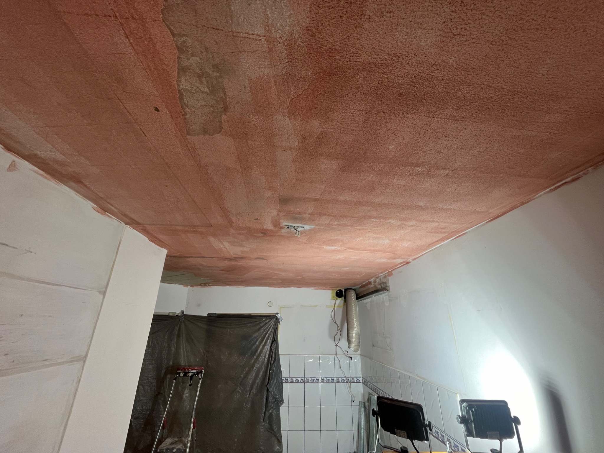 Czerwona warstwa gruntująca na suficie w trakcie remontu. Widoczne ślady po szpachlowaniu, drabina i lampy budowlane. Przygotowanie do malowania sufitu.