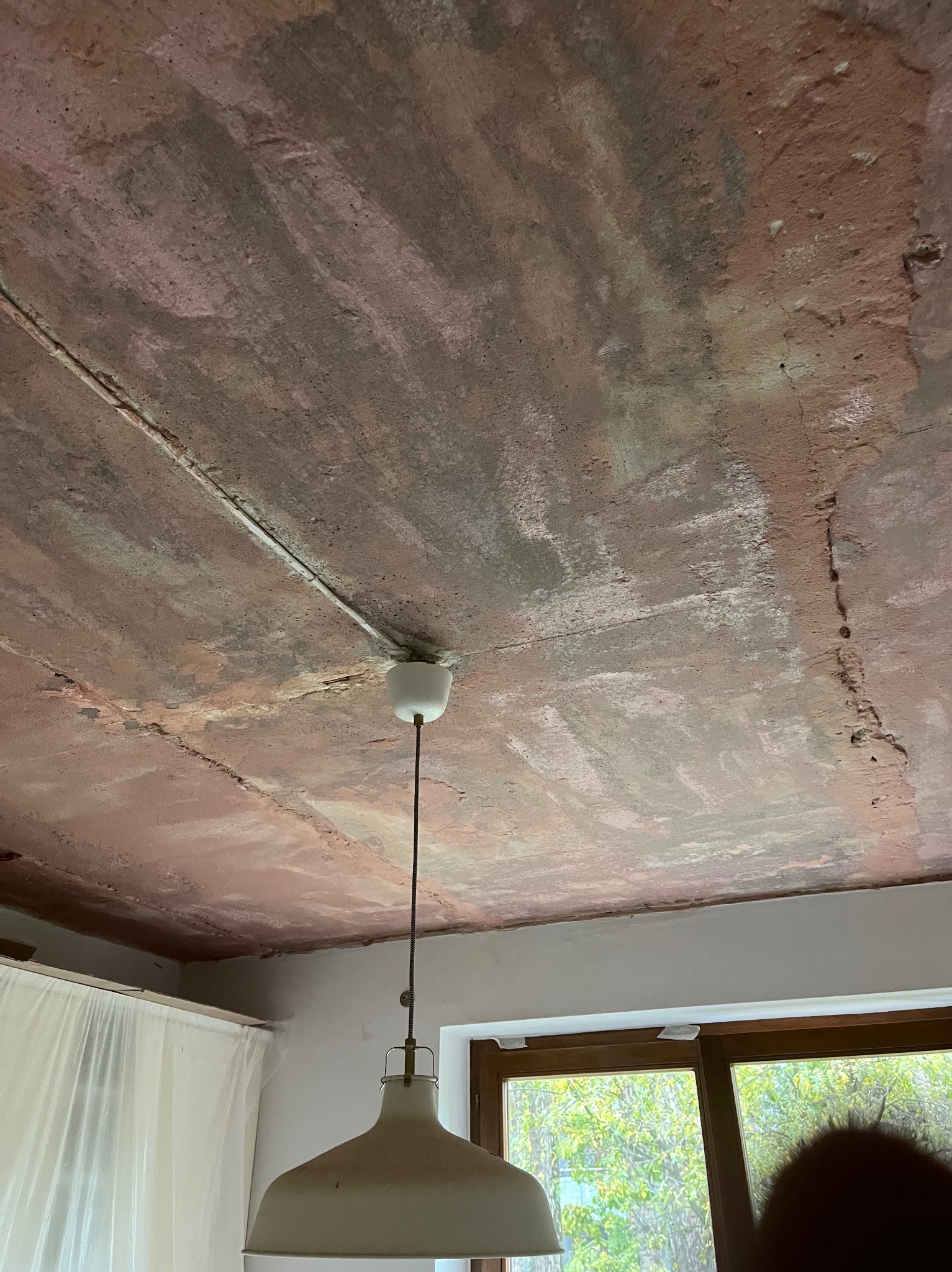 Niedokończony sufit z widocznymi śladami po szpachlowaniu i instalacji elektrycznej, z lampą wiszącą na środku. Widoczne okno i zasłonięty fragment ściany.