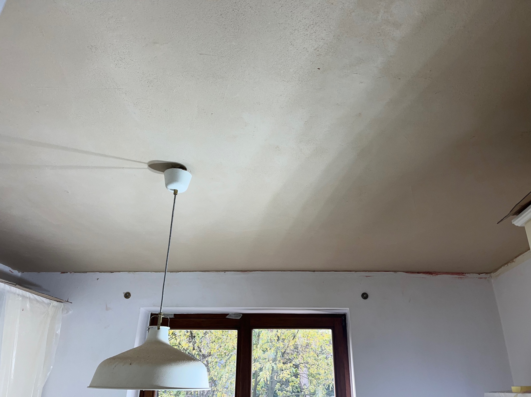 Wnętrze pokoju w trakcie szpachlowania sufitu, widoczna lampa wisząca i okno z widokiem na drzewa. Ślady szpachli na suficie i ścianach.