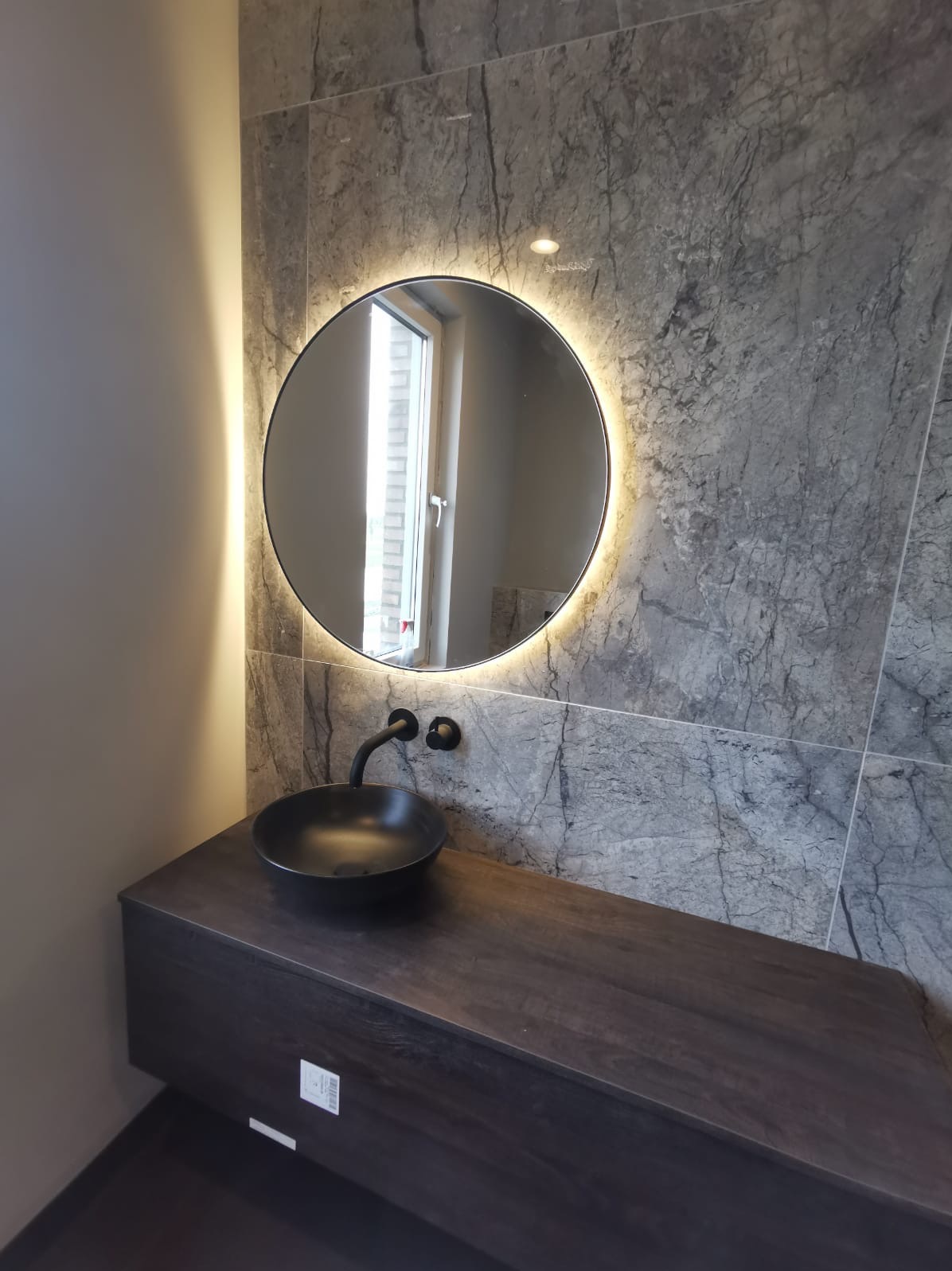 Nowoczesna łazienka z okrągłym lustrem LED, czarną umywalką nablatową i szarą marmurową ścianą. Minimalistyczny design, eleganckie wykończenie.