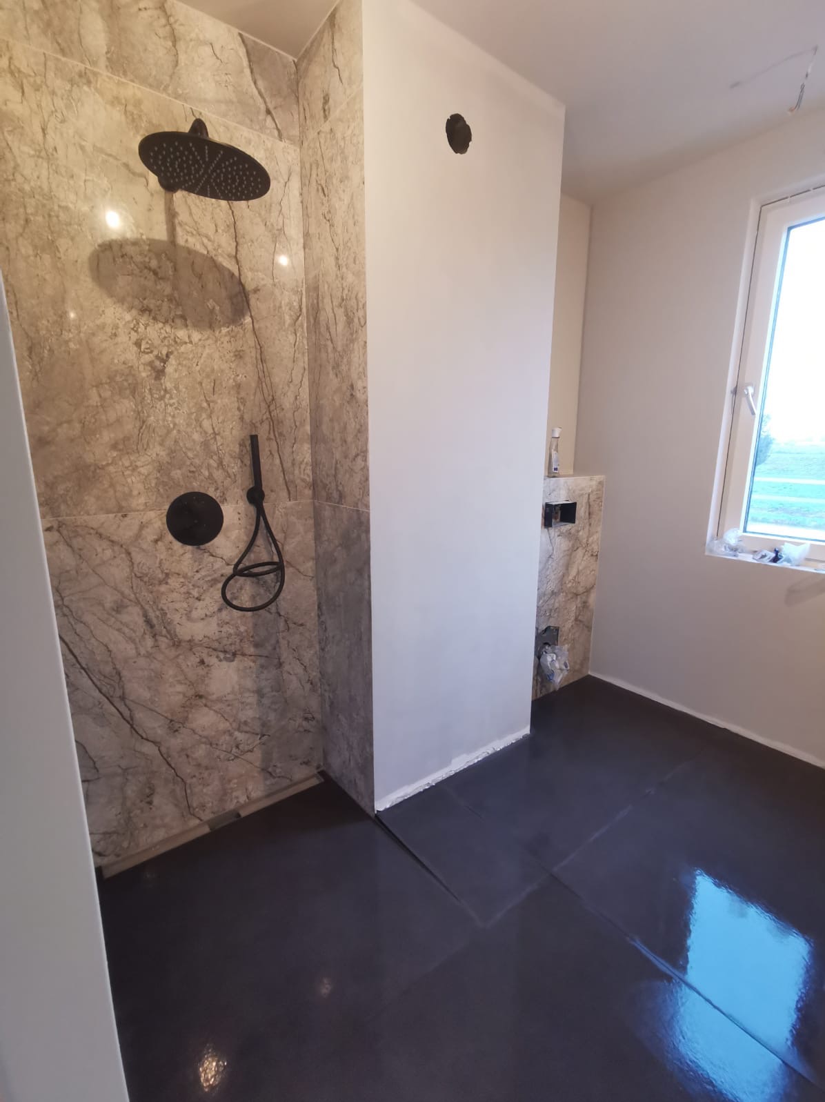 Wykończona łazienka z marmurowymi płytkami pod prysznicem i czarną podłogą. Widoczna armatura w kolorze czarnym i okno z widokiem na zewnątrz.