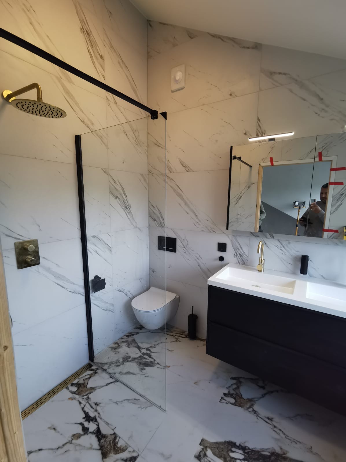 Nowoczesna łazienka z marmurowymi płytkami, czarnymi akcentami i złotą armaturą. Kabina prysznicowa typu walk-in z czarną ramą. Umywalka z szafką w kolorze czarnym.