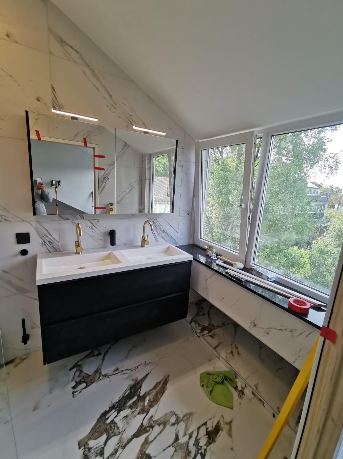 Wykończona łazienka z marmurowymi płytkami, czarną szafką pod umywalką z dwiema białymi umywalkami i złotą armaturą. Widok na zieleń za oknem.