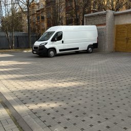 MATBUS Mateusz Stefaniak - Biały bus dostawczy zaparkowany na brukowanym placu, budynek w tle. Dzień, słonecznie, widok z boku. Charakterystyczny wzór kostki brukowej.