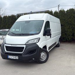 MATBUS Mateusz Stefaniak - Biały bus Peugeot na parkingu, z widoczną tablicą rejestracyjną i naklejką z imieniem kierowcy na szybie. W tle inne pojazdy i rząd zielonych krzewów.