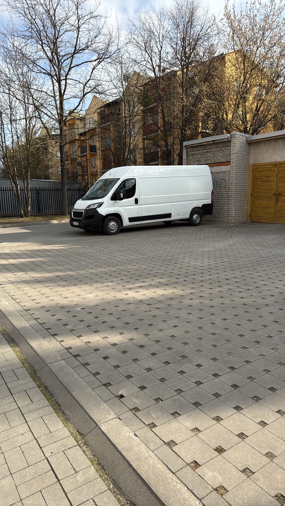 Biały bus dostawczy zaparkowany na brukowanym placu, budynek w tle. Dzień, słonecznie, widok z boku. Charakterystyczny wzór kostki brukowej.