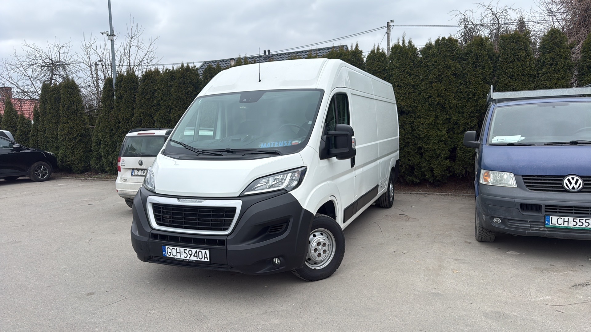 Biały bus Peugeot na parkingu, z widoczną tablicą rejestracyjną i naklejką z imieniem kierowcy na szybie. W tle inne pojazdy i rząd zielonych krzewów.