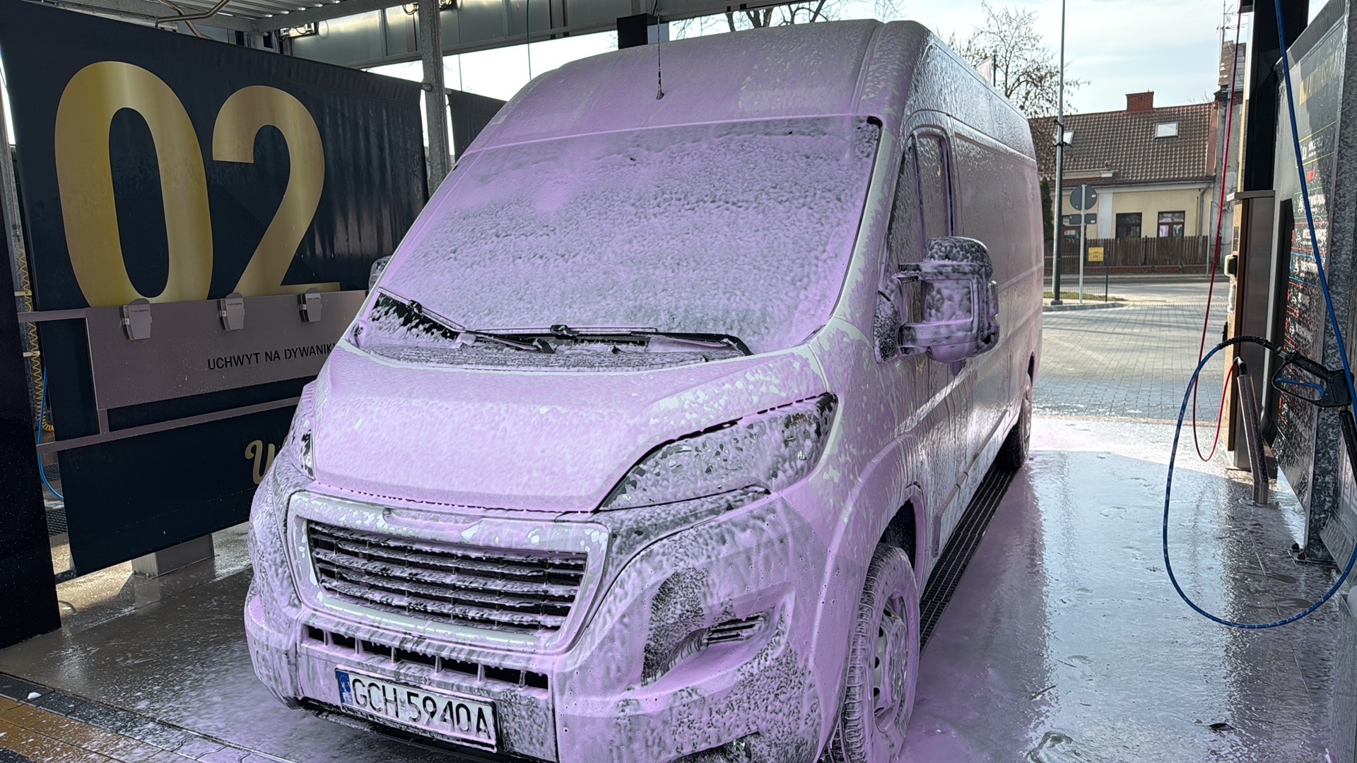 Bus w pianie podczas mycia w samoobsługowej myjni. Widoczny numer stanowiska '02' i uchwyt na dywaniki. Piana pokrywa całą karoserię, tablica rejestracyjna widoczna.