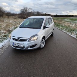 Detailing84 - Srebrna Opel Zafira na mokrej, asfaltowej drodze w zimowym krajobrazie. Widoczne włączone światła mijania i tablica rejestracyjna GWE 5222V.