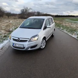Srebrna Opel Zafira na mokrej, asfaltowej drodze w zimowym krajobrazie. Widoczne włączone światła mijania i tablica rejestracyjna GWE 5222V.