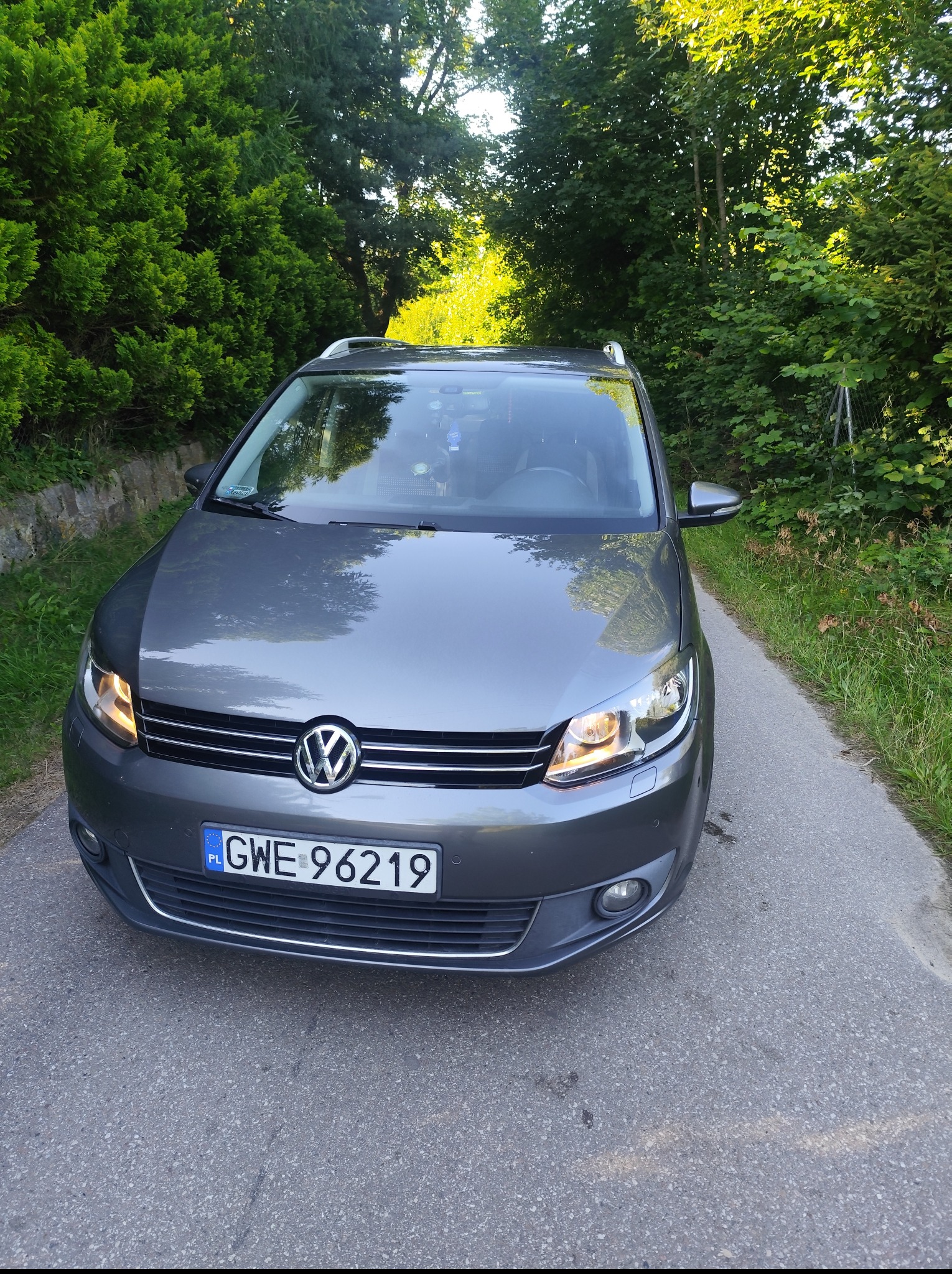 Szary Volkswagen Touran na asfaltowej drodze, otoczony zielenią. Widoczne włączone światła i polska tablica rejestracyjna GWE 96219.